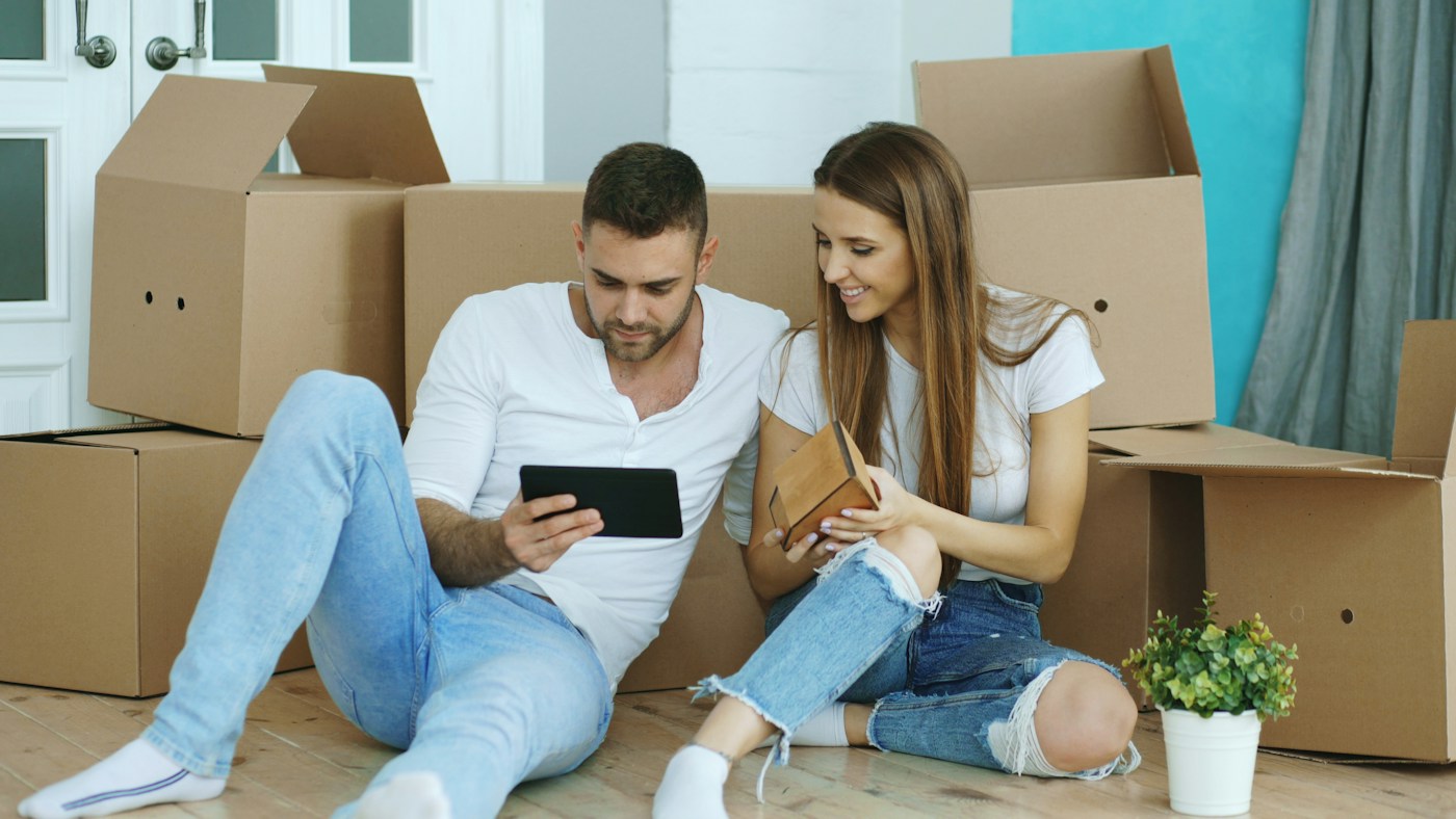 Two renters sitting on the floor among moving boxes, checking something on a tablet